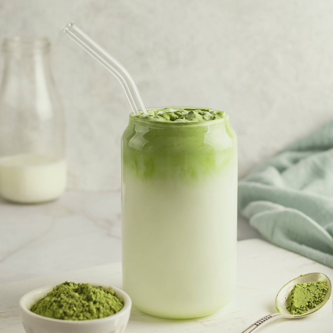 Creamy matcha latte in glass jar with straw, fresh matcha powder in bowl and spoon, milk bottle background